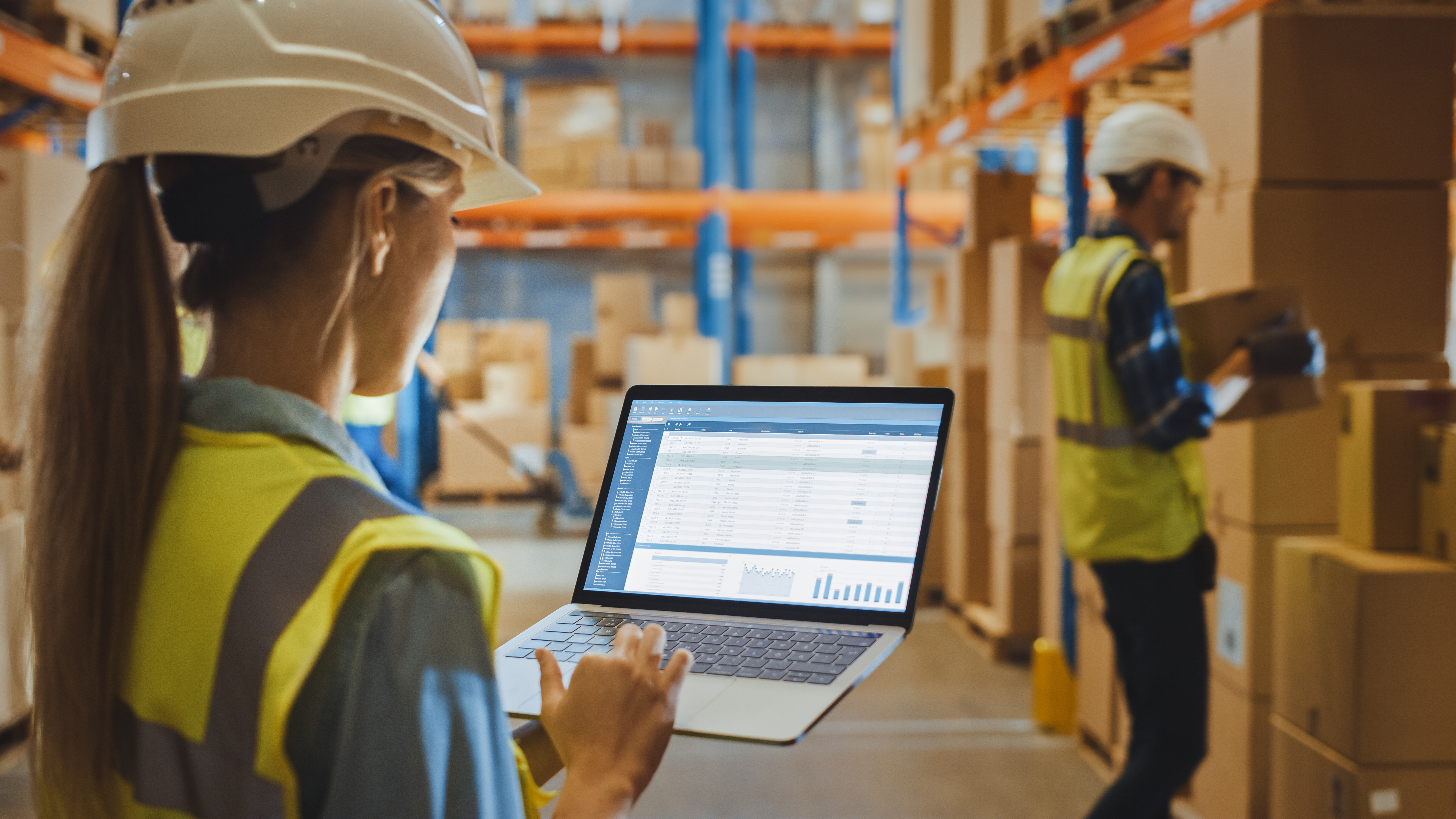 Kvinnlig lagerarbetare med hjälm granskar en rapport om Warehouse Profitability på sin laptop.